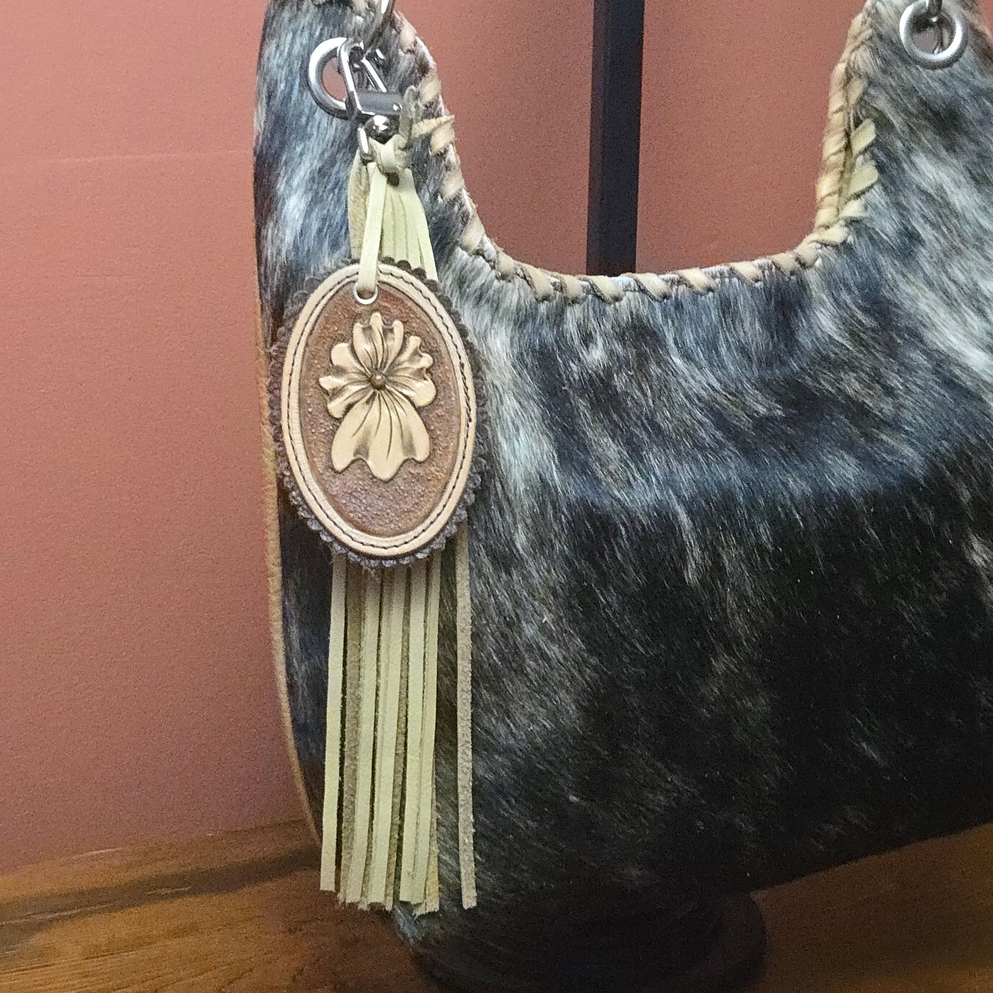 A close-up of a shoulder bag made from black and gray mottled cowhide, featuring a cream-colored leather tassel charm with a carved floral medallion, displayed on a stand against a dusty terracotta background.