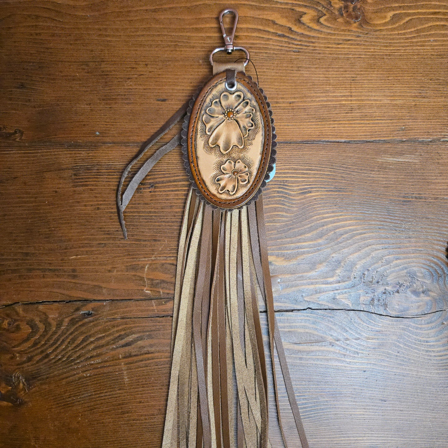 A brown leather fringe keychain or bag charm with an oval tooled leather center featuring floral designs, resting on a rustic wooden surface.
