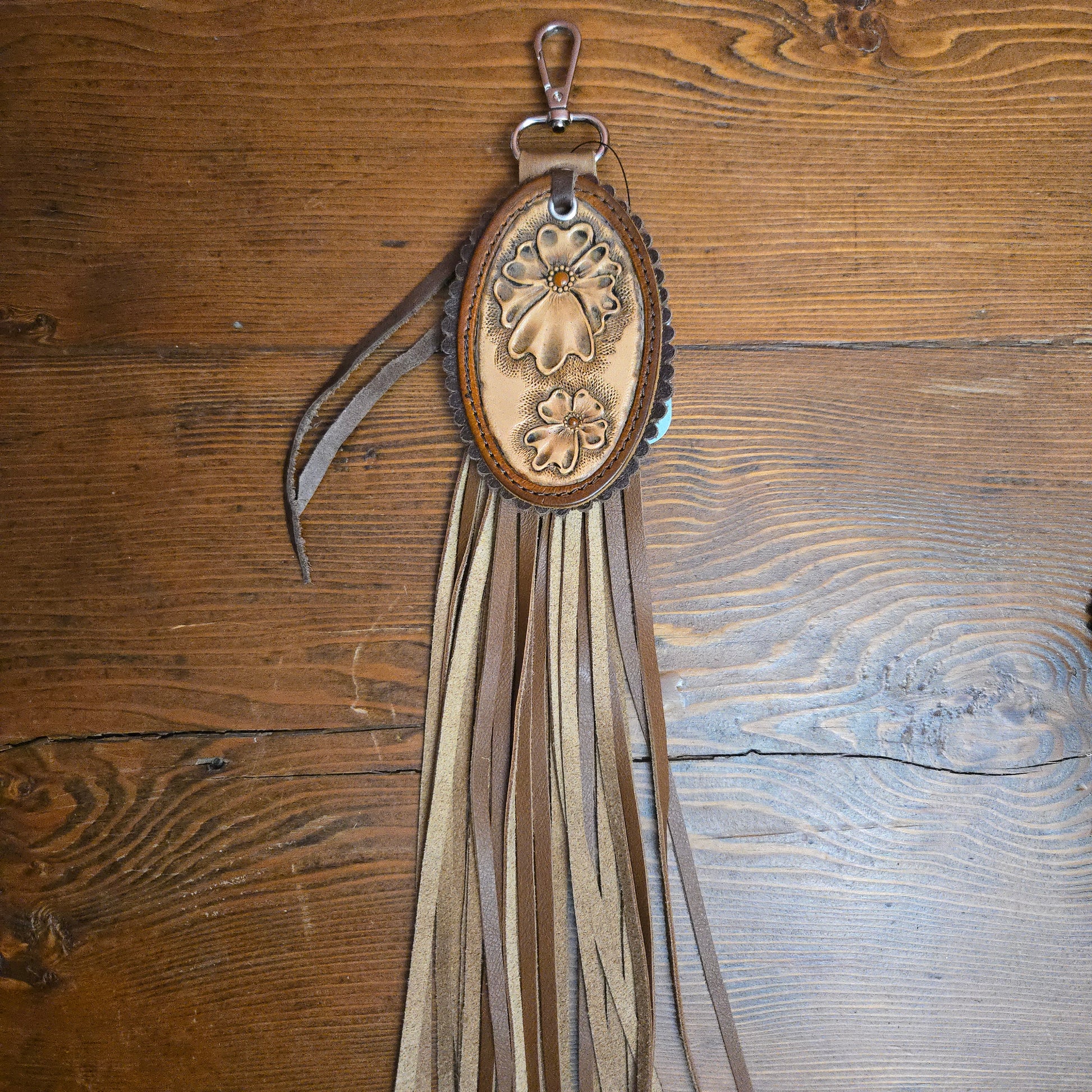 A brown leather fringe keychain or bag charm with an oval tooled leather center featuring floral designs, resting on a rustic wooden surface.