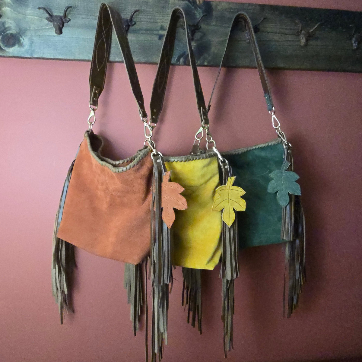 Three fringe shoulder bags in brown, mustard yellow, and dark green hang from a rustic wooden rack against a dusty rose wall. Each bag has a wide body, long leather straps, and a leaf-shaped charm in a matching color.