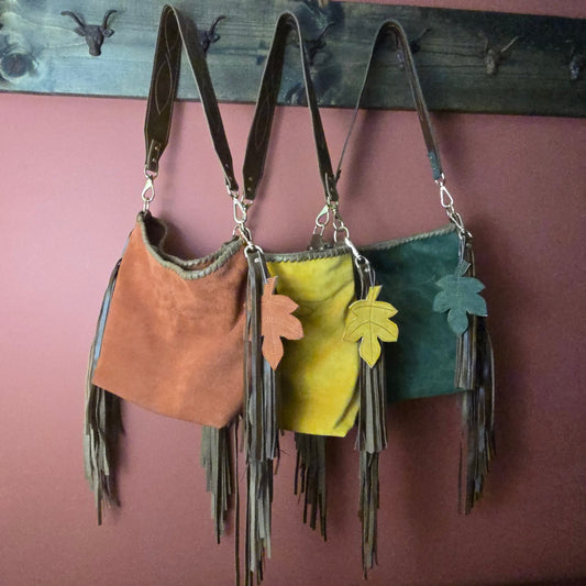 Three fringe shoulder bags in brown, mustard yellow, and dark green hang from a rustic wooden rack against a dusty rose wall. Each bag has a wide body, long leather straps, and a leaf-shaped charm in a matching color.