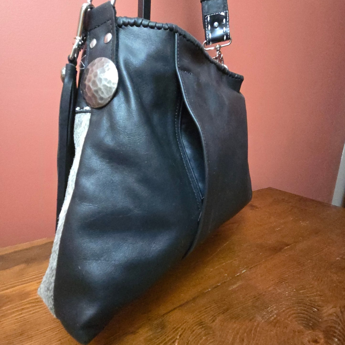 A black leather shoulder bag with silver-tone hardware, including a hammered metal concho, resting on a wooden table against a terracotta-colored wall.