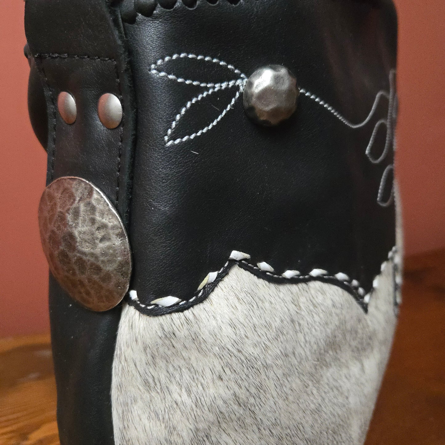 Close-up of a western-style  bag featuring black leather, white hair-on-hide, silver conchos, white stitching, and a warm terracotta background.