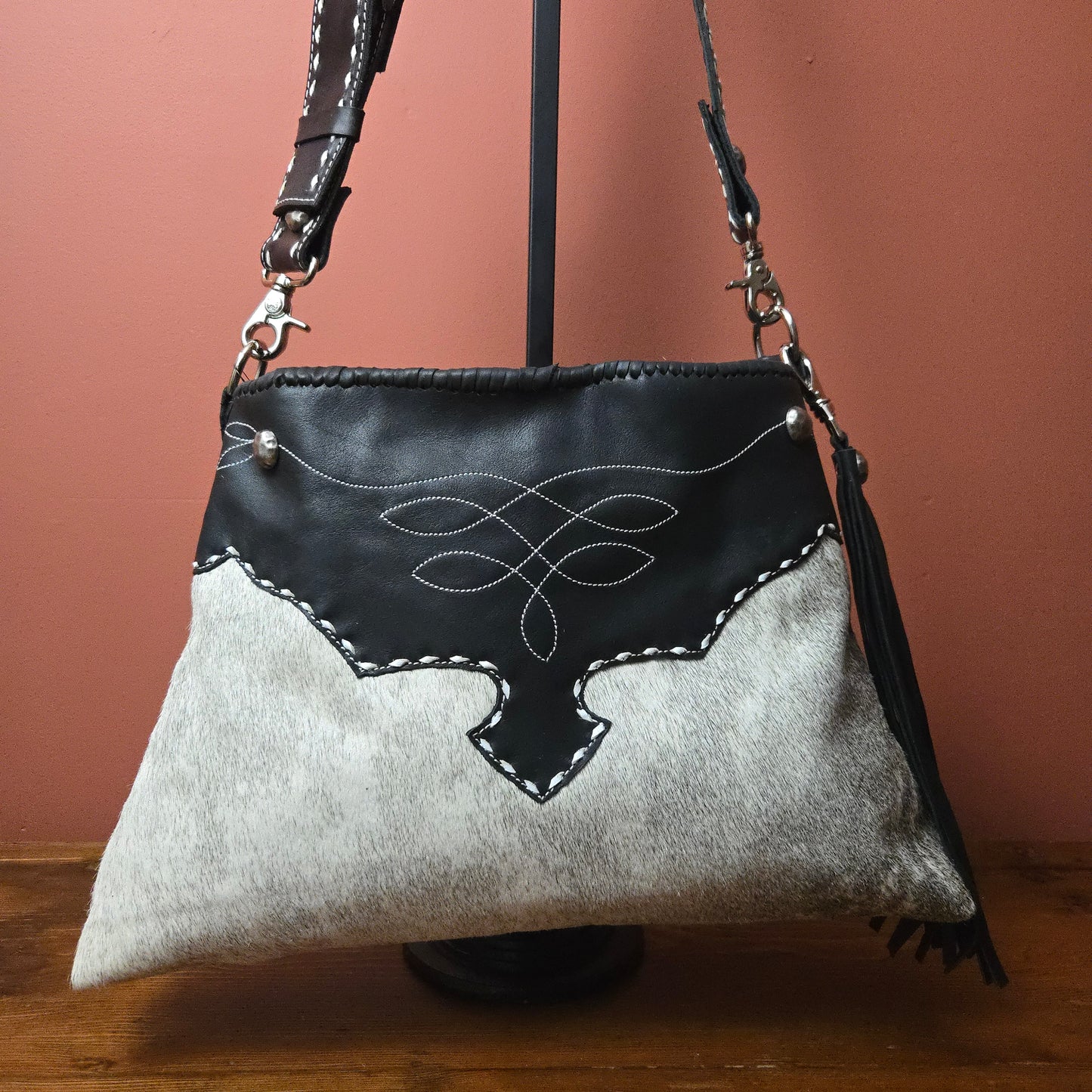 A western-style shoulder bag made from black leather and light gray cowhide, featuring white contrast stitching, decorative stitching on the front, and a black leather tassel, displayed on a black stand against a terracotta background.