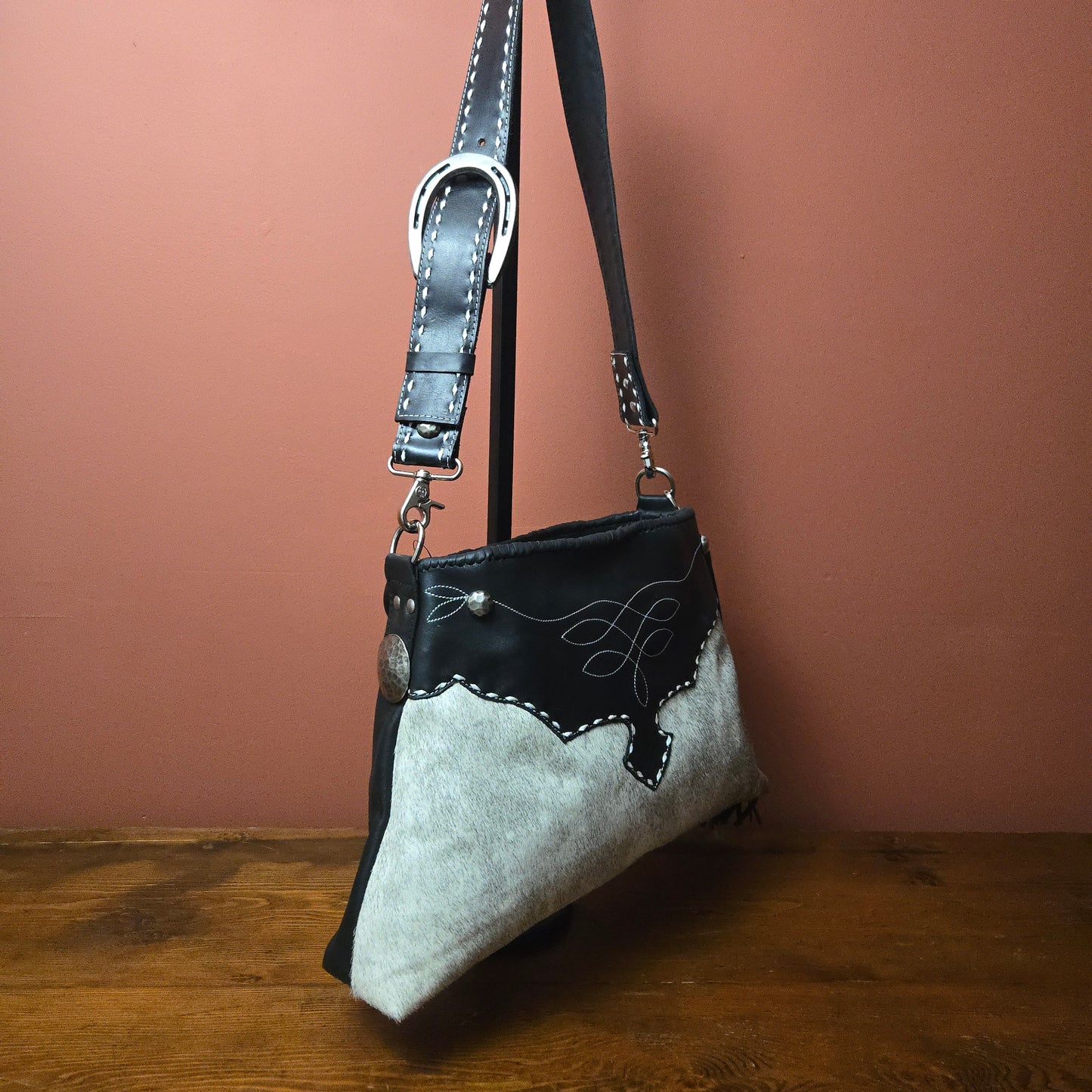 A western-style shoulder bag made from black leather and Hair on Hide, featuring silver-tone hardware, Buckstitch, and a Hammered Nickel  concho accent, hanging against a terracotta-colored wall on a wooden surface.