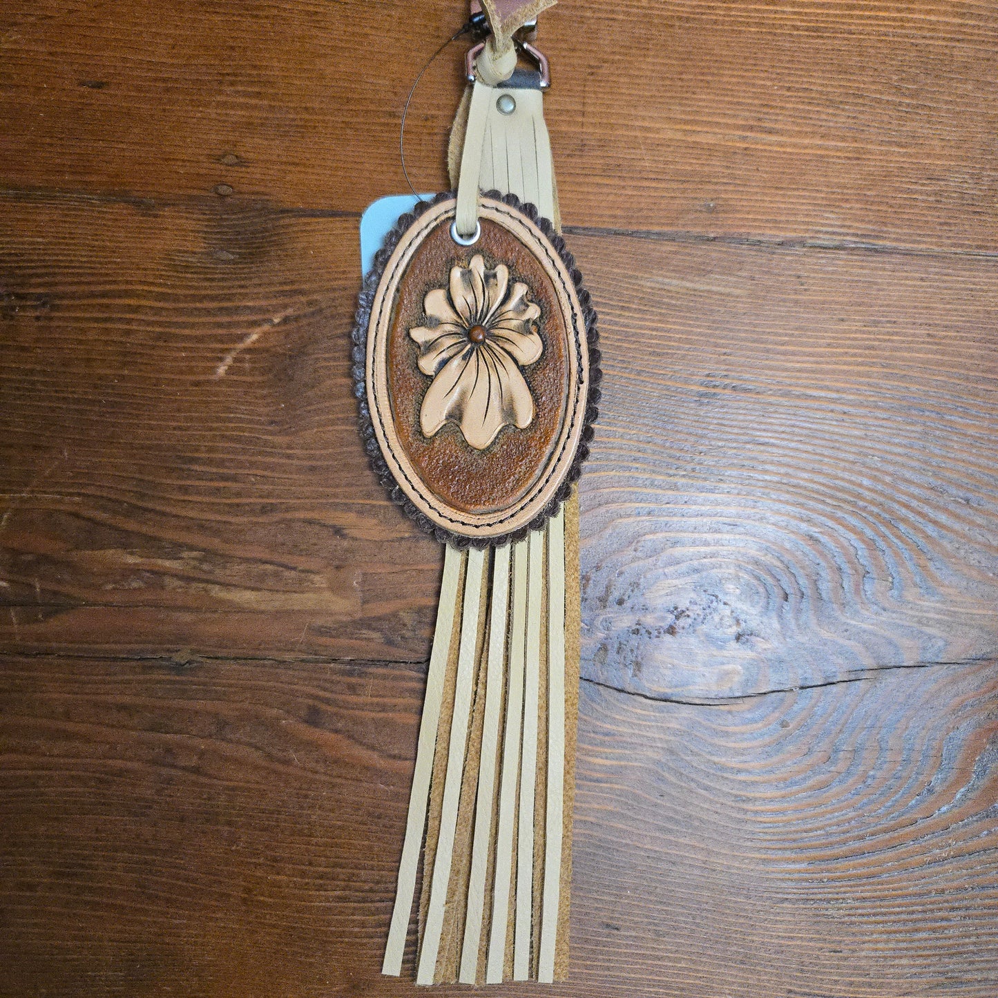 A western-style leather Keychain or bag charm with an oval tooled leather center featuring a floral design, fringe tassel, photographed on a wooden surface.
