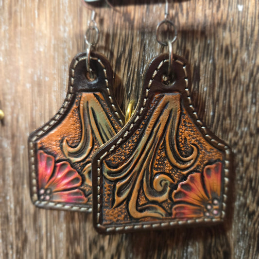Tooled leather earrings with floral designs, featuring brown, gold, and red accents on a rustic wooden background.
