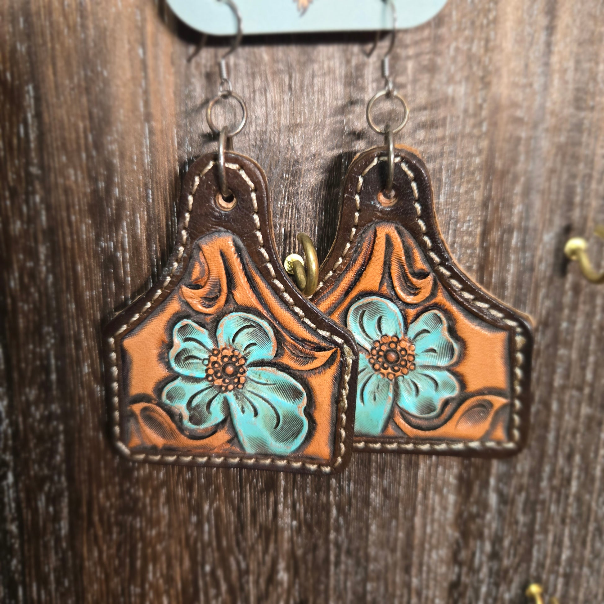 A pair of tooled leather western-style earrings with turquoise floral designs, displayed on a dark wood background.