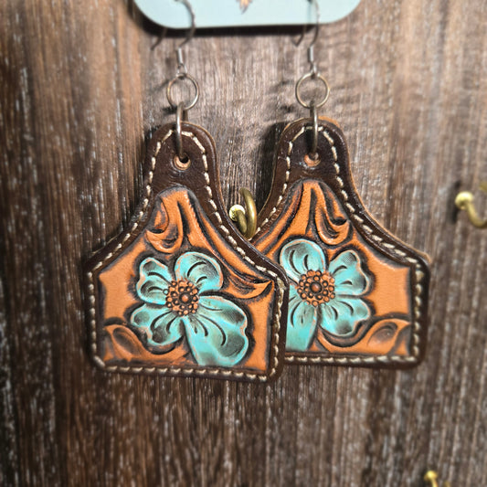 A pair of tooled leather western-style earrings with turquoise floral designs, displayed on a dark wood background.